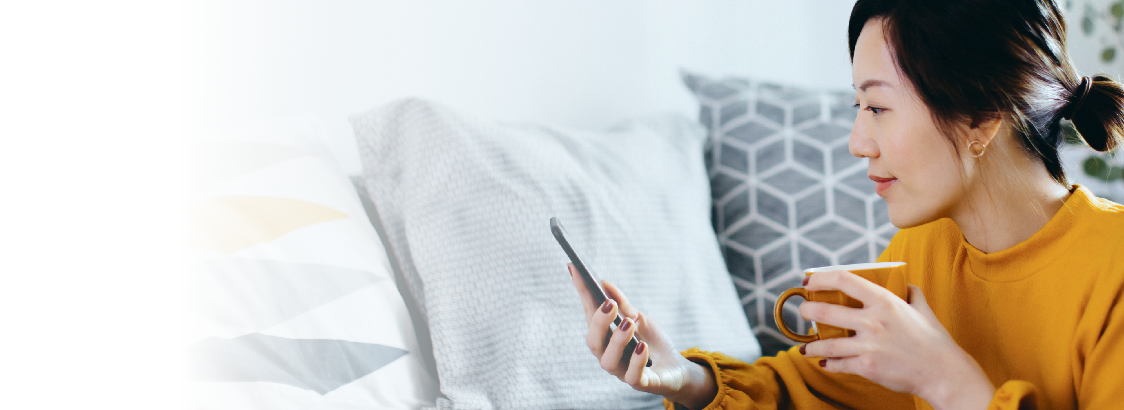 Beautiful smiling young Asian woman chilling at cozy home, sitting on the floor by the sofa, enjoying a cup of coffee and using smartphone