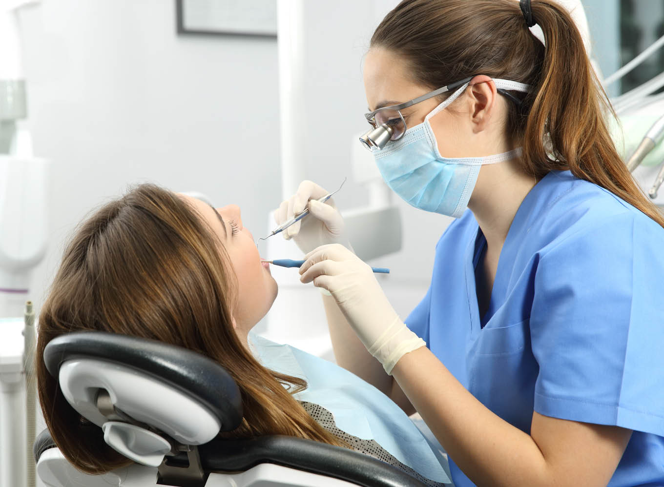 Dentist wearing eyeglasses gloves and mask examining a patient teeth with a dental probe and a mirror in a clinic box with equipment in the background