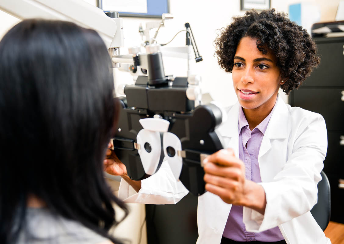 A African American optometrist is checking her patient's eye sight. Optometrist is ethnic of African Decent. Doctor is in ethnic minority. She is checking her patient's eye sight by pulling eye test equipment - Phoropter. Its real optometrist office.   
