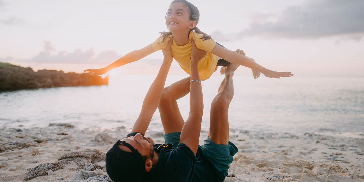 Parent and child play on the beach at sunset, with the child held in the air like an airplane, smiling.
