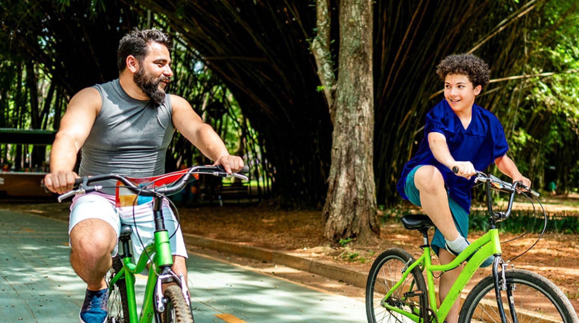Father and son ride bikes together on a tree-lined path, smiling at each other.