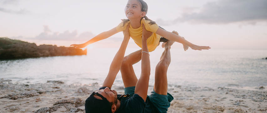 Parent and child play on the beach at sunset, with the child held in the air like an airplane, smiling.