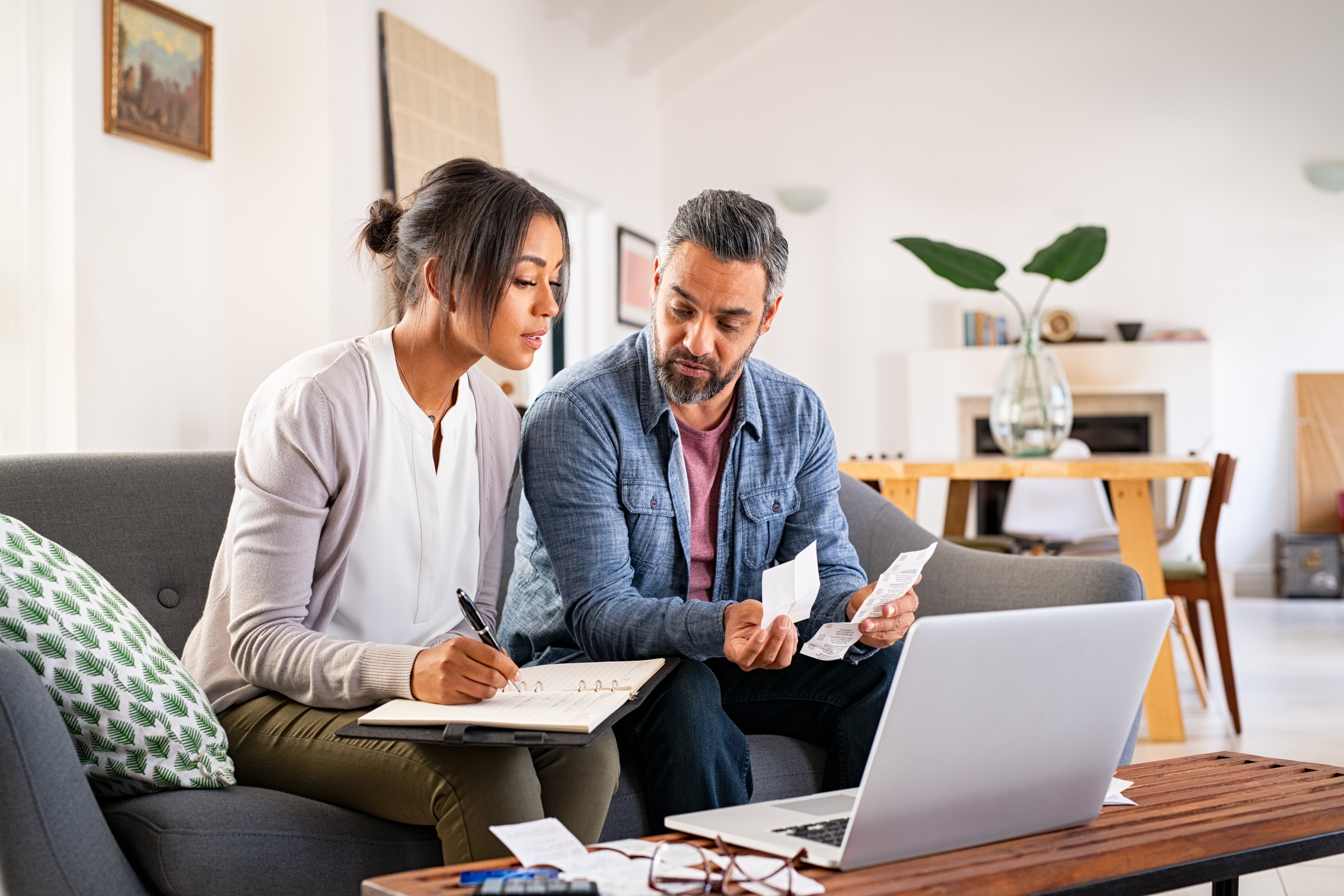 Couple looks through paper bills together in front of a laptop screen. 