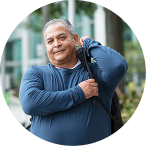 A mature Hispanic man, in his 50s, walking in the city carrying a backpack. He is looking at the camera with a serious expression.